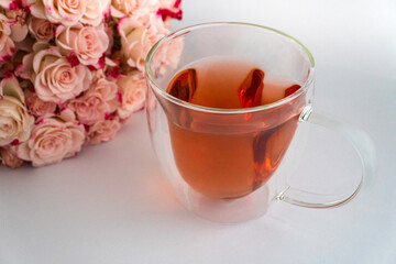 Herzförmige Teetasse aus Glas mit rotem Tee gefüllt auf weißem Satinstoff, hellrosafarbene Rosen oben links, Schrägansicht, horizontal 