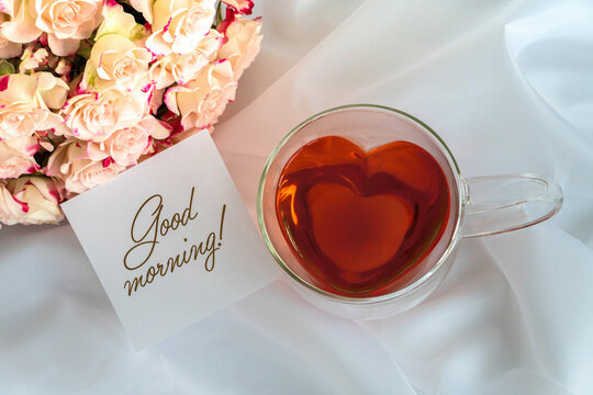 Herzförmige Teetasse aus Glas mit rotem Tee gefüllt auf weißem Satinstoff, hellrosafarbene Rosen oben links und ein weißer Notizzettel mit dem Schriftzug Good morning im Bild, Topview, horizontal