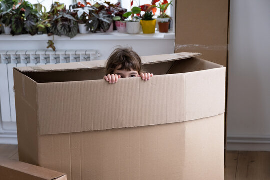 The Girl Hides In A Big Box And Looks Out Of It Funny In A Room Inside The Home. Moving To A New House, Unpacking Things, Renting An Apartment, Mortgage, Housing Issue