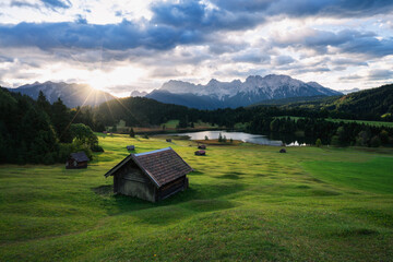 Sonnenaufgang mit Hütte, See, Bergen und grüner Wiese mit weiß blauem Himmel.