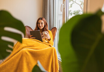 Young attractive redhead woman holding a tablet at home, with a blanket in the armchair by the...