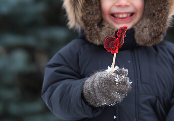 Portrait of a beautiful little Russian boy in a hat with earflaps with a lollipop cockerel in...