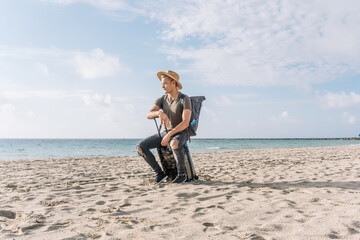 portrait of a lost man sitting on his small suitcase and looking at the sea on the beach,