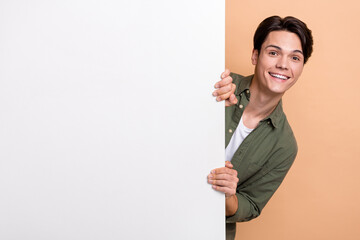 Portrait of young funny excited guy wearing khaki shirt hiding behind huge whiteboard promo...