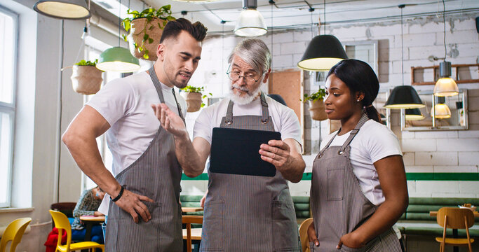 Portrait Of Serious Caucasian Old Bearded Man In Own Restaurant In Apron Tapping On Tablet Looking At Screen Discussing With Mixed-race Young Male And Female Workers Colleagues Something. Cafe Concept