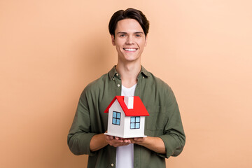 Portrait of satisfied cool cute man with brunet haircut dressed khaki shirt holding small house isolated on beige color background