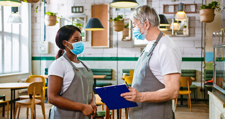 Portrait of mixed-race workers in cafe at work. Old man measuring temperature of young African American female employee in medical mask and apron using infrared thermometer on forehead, health check
