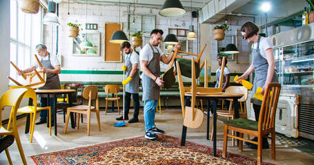 Mixed-race different people workers in coffee house working together putting chairs on tables to...