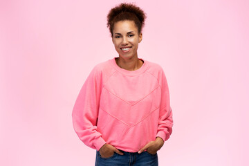 Young cute woman with afro hair, holding hands on jeans pockets, smiling at camera, pink background