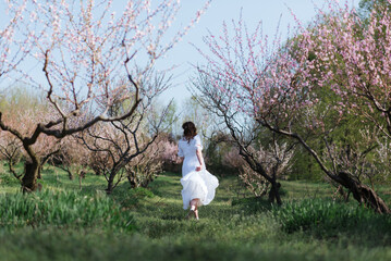 Young girl in white dress running in blooming spring peach tree garden