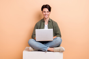 Full length cadre of young optimistic man sitting platform cube use laptop browsing information...