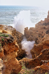 Wave splashing over orange rocks in the Atlantic Ocean on a warm winter day in Faro, Portugal near a beach in Carvoeiro.