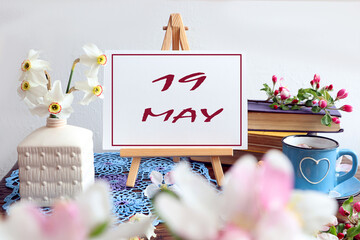 Calendar for May 19: the name of the month May in English, the numbers 19 written on paper, among the flowers of the apple tree lying on the books, a bouquet of daffodils in a white vase