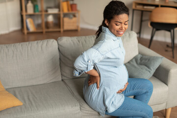 Sad black pregnant lady with big belly suffering from back pain, sitting on sofa in living room,...