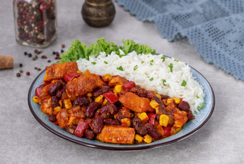 Chicken and tomato ragout with red beans, corn and bell pepper served on a plate with portion of cooked white rice. Selective focus, gray concrete background. Horizontal.