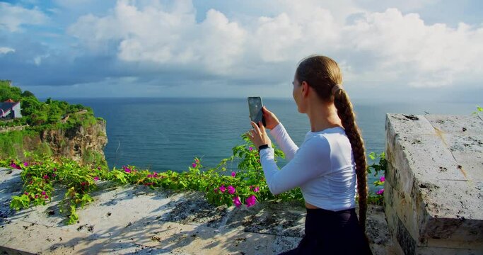 Girl taking pictures of ocean on phone in Uluwatu Hindu Temple Bali Island Indonesia. Woman tourist using smartphone and relaxing at rocky beach. Limestone sheer cliffs and azure Indian waters.
