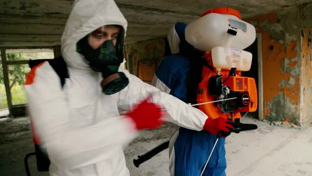 Epidemiologist in protective suit starts motor of disinfection sprayer device on coleague back among abandoned building closeup