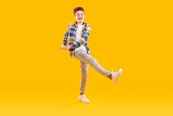 Fototapeta na wymiar Happy funny redhead teenage boy raised up his leg. Full length shot of attractive joyful boy in casual plaid shirt and jeans standing on one leg over isolated yellow studio background