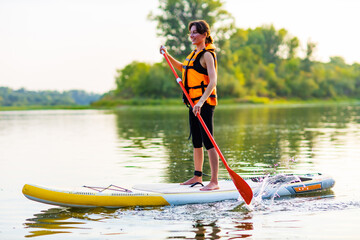 young woman in orange life jacket on supboard at river