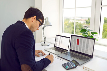 Fototapeta na wymiar Financial accountant working in modern office. Busy man in suit and glasses sitting at desk by window, using calculator and two laptop computers, working with digital sheets and taking notes on paper