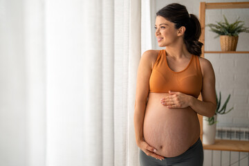 Happy pregnant woman standing by the window, wearing an orange sports bra and gray leggings is smiling and touching her stomach.