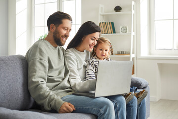 Happy family of mom, dad and cute little daughter sitting at laptop at home. Young parents and toddler kid having video call via laptop at home. Family sitting on sofa using modern device