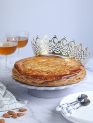 A traditional French galette des rois or king cake on a plate with crown and glasses of cider in background with spoon. Cake made with puff pastry and creamy almond filling roll in a circle shape