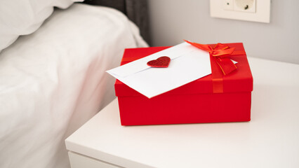 Romantic love letter in white envelope with Valentines heart and gift box with red ribbon on white bedside table. Happy Valentine's Day concept
