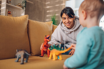 Mother and toddler son playing with dinosaurs toys at home on the couch - obrazy, fototapety, plakaty