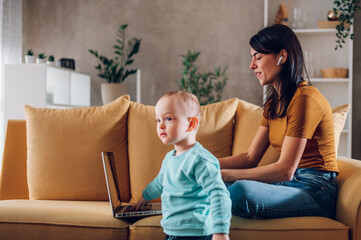 Mother playing and hugging with her child son while using laptop at home