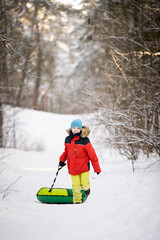 Cute happy baby runs through the winter forest and carries a snow inflatable sled. Baby in bright overalls in a snowy winter forest. Activities for children