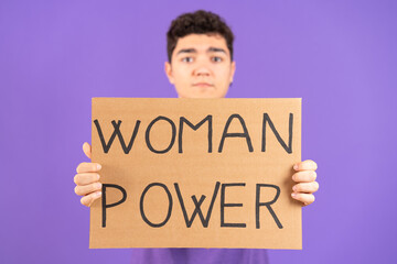 Hispanic teenager boy showing cardboard with feminist message isolated on purple background. Feminism and woman power concept