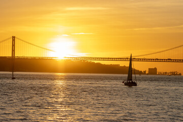 Fototapeta premium Bright orange sunset over the river and a sailing boat floating towards the bridge