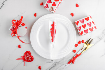 Valentines day dinner background with gifts, wine glass with red hearts, plate, cutlery on white marble. Romantic holiday table setting. Festive table setting on marble background. Top view, flat lay