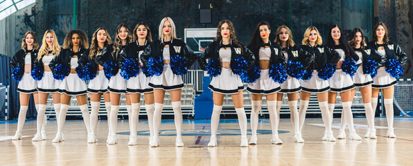 Front view of cheerleaders in uniforms standing in row and holding blue shiny pom-poms. Basketball...