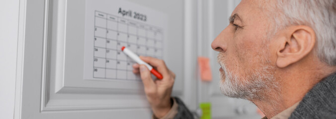 side view of senior man with alzheimer disease pointing with felt pen at calendar in kitchen, banner.