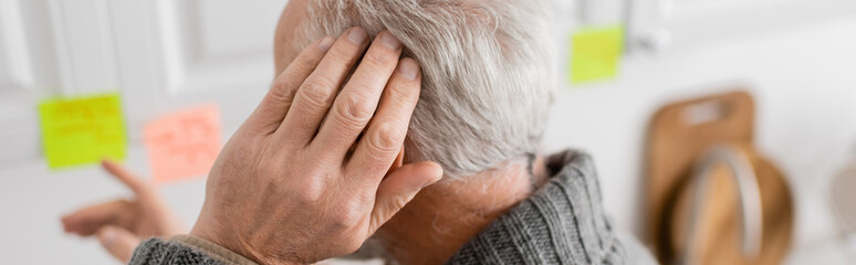 Fototapeta premium aged man with alzheimer disease touching head and pointing at blurred sticky notes in kitchen, banner.