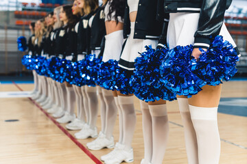 Close up shot of cheerleaders standing in line on basketball court and holding blue pom-poms. High...