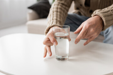partial view of aged man with parkinson disease and trembling hands taking glass of water from table at home.