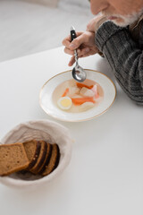 partial view of aged man suffering from parkinson disease and sitting with spoon near soup and bread in kitchen.