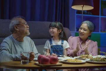 Happy Asian family grandmother grandfather and granddaughter dining on table and having fun during at home night time, senior parent and child eating food together in living room indoors together