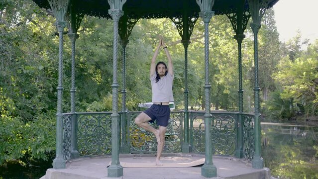 Traveling in of a young latin hippie man with long hair starting the yoga pose vrikshasana on a gazebo in front of a lake. 4k video.