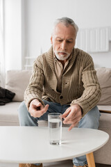 senior grey haired man with parkinson disease and tremor in hands sitting near glass of water on...