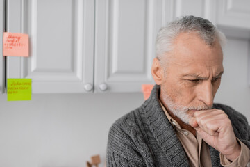 Fototapeta premium aged man with alzheimer syndrome holding fist near face while thinking near blurred sticky notes in kitchen.