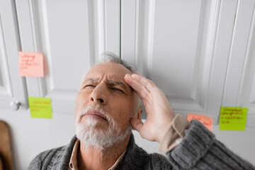 senior man with closed eyes and hand near head standing near blurred sticky notes while suffering...