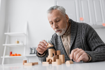 bearded senior man suffering from alzheimer disease and playing building blocks game on table at home.