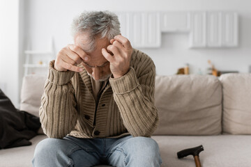 senior man with parkinson disease and trembling hands sitting with bowed head on couch at home.