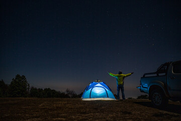 Fototapeta na wymiar Night camping. Man hikers stands and raise both arms up from enjoying the beautiful night sky with stars. Male trekkers relaxing and enjoying mountain camping in nature.