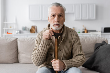 depressed man suffering from dementia while sitting with walking cane and looking at camera at home.