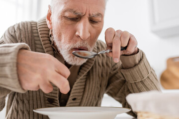 aged man suffering from parkinsonian syndrome holding spoon in trembling hand while having dinner at home.